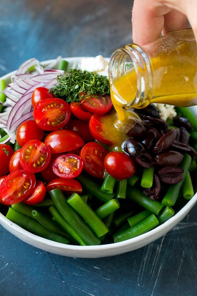 Salad dressing being poured over green beans, cherry tomatoes, olives, herbs and onions.