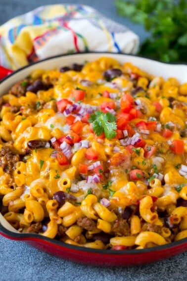 A skillet of chili mac topped with melted cheese, diced tomatoes and minced onion.