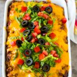 Taco casserole in a baking dish topped with melted cheese, lettuce, tomato and olives.