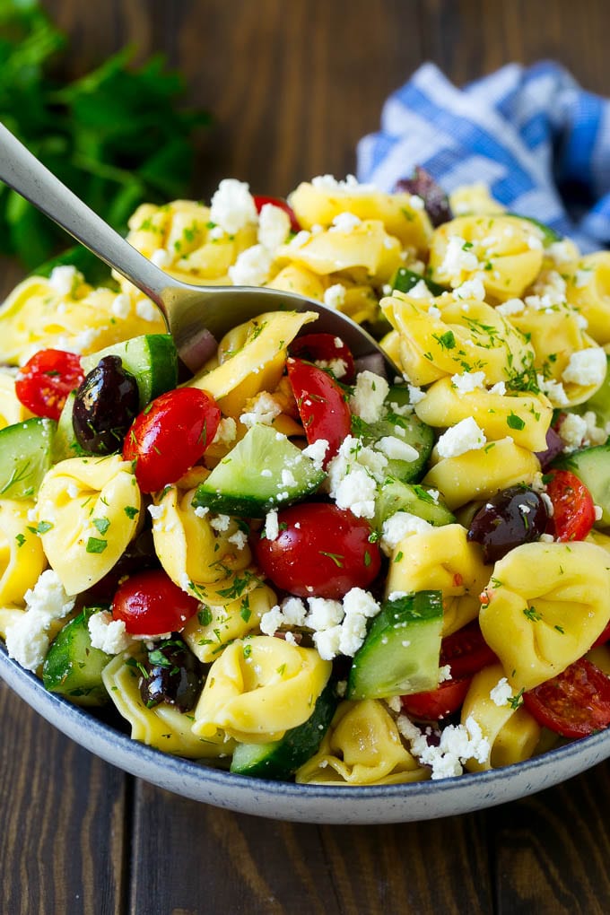A bowl of Greek tortellini salad with a serving spoon in it.