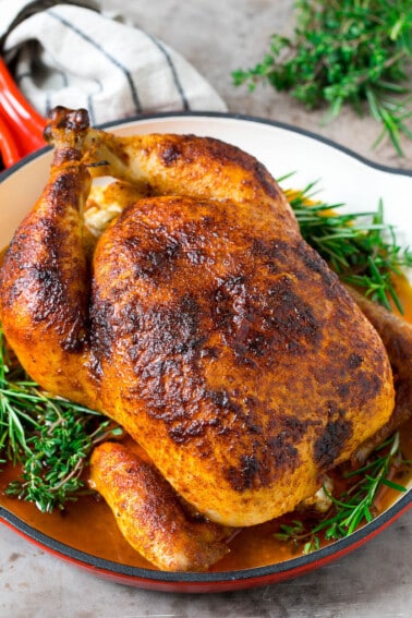 A rotisserie chicken in a red skillet with sprigs of fresh herbs.