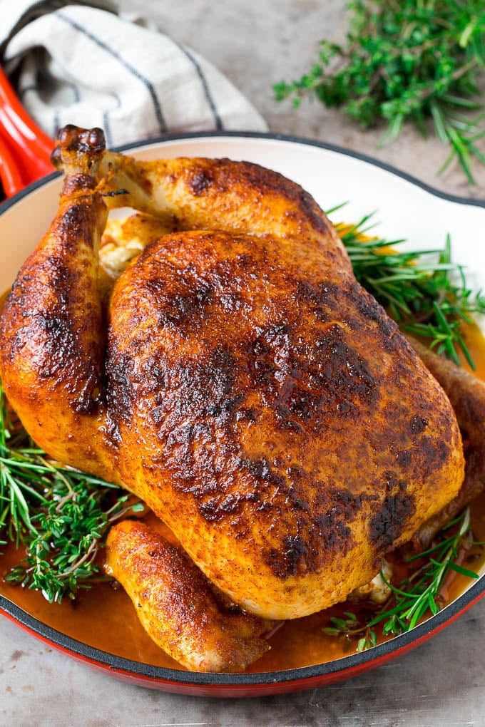 A rotisserie chicken in a red skillet with sprigs of fresh herbs.