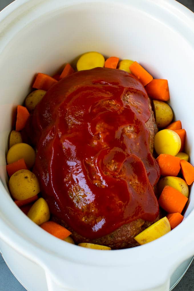 A loaf of ground beef topped with ketchup in a slow cooker, surrounded by carrots and potatoes.