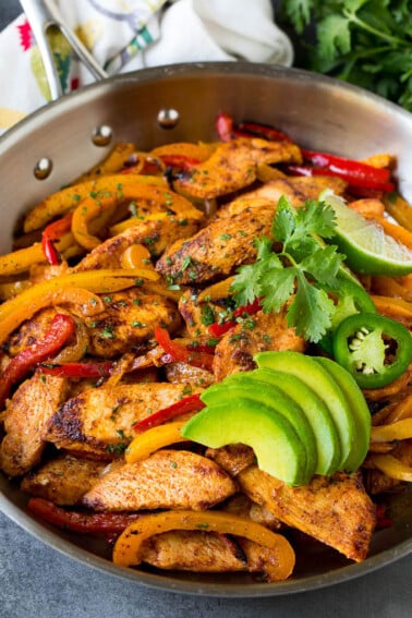 A pan of chicken fajitas with peppers and onions, topped with sliced avocado and cilantro.