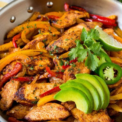 A pan of chicken fajitas with peppers and onions, topped with sliced avocado and cilantro.