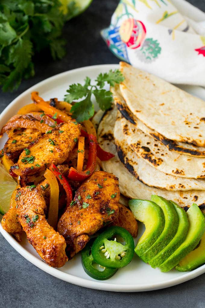 A plate of chicken fajitas with sliced avocado, tortillas and herbs.