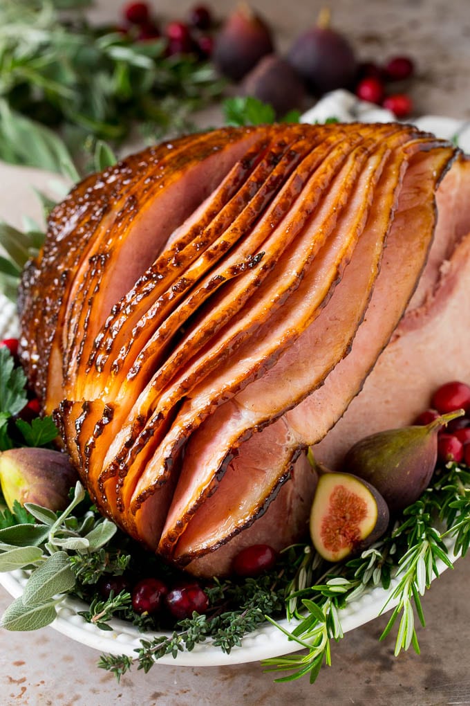 A honey glazed ham on a serving platter, decorated with fruit and herbs.