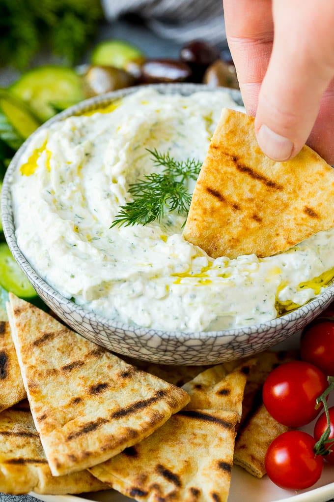 The best homemade tzatziki sauce with cucumber and herbs mixed into Greek yogurt. #appetizer #dinneratthezoo A hand dipping pita bread into a bowl of tzatziki.