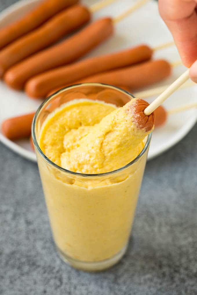 A hot dog being dipped into a cup of corn batter.
