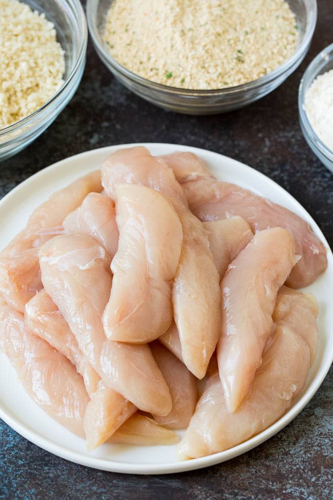 Raw chicken pieces on a plate with bowls of breadcrumbs on the side.