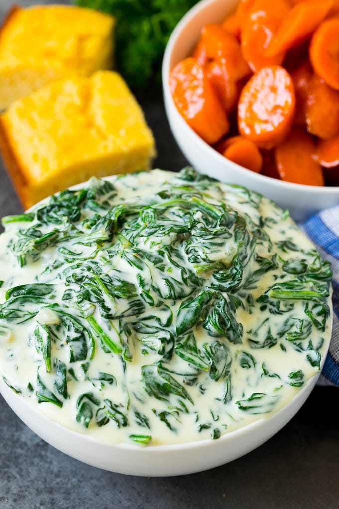 A bowl of creamed spinach next to a bowl of glazed carrots and cornbread.