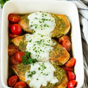 Pesto chicken in a baking dish with melted cheese and cherry tomatoes.