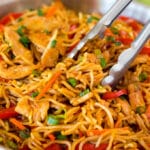 Tongs serving up a portion of yakisoba noodles with vegetables and chicken.