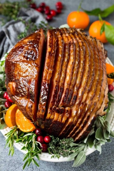 Baked ham coated in glaze, served on a platter with herbs and fruit.