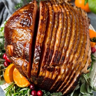 Baked ham coated in glaze, served on a platter with herbs and fruit.