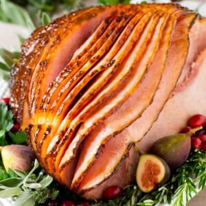 A honey glazed ham on a serving platter garnished with herbs and fruit.