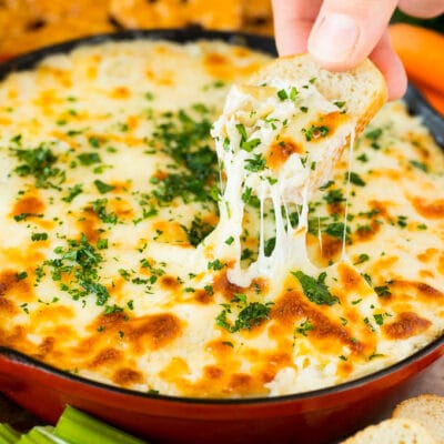 A skillet of artichoke dip with a hand reaching to take a serving.