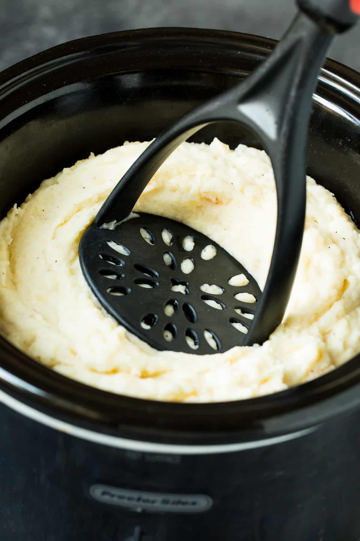 A masher in a slow cooker full of potatoes.