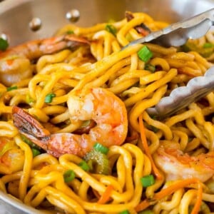 A pan of shrimp lo mein with tongs serving up a portion.