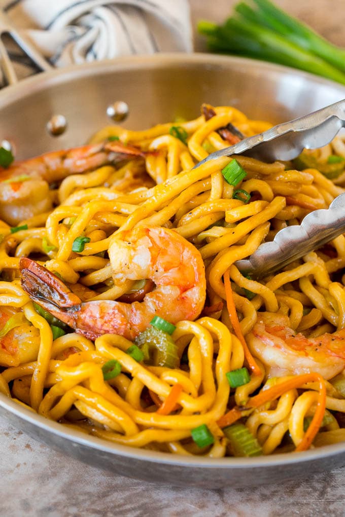 A pan of shrimp lo mein with tongs serving up a portion.