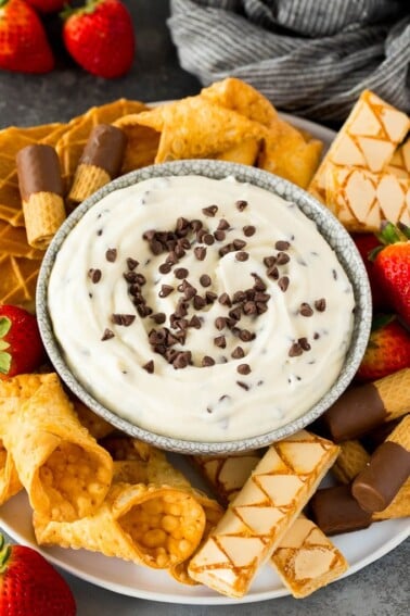 A bowl of cannoli dip surrounded by cookies, cannoli shells and strawberries.