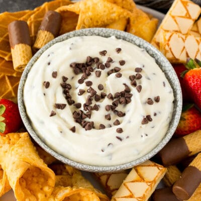 A bowl of cannoli dip surrounded by cookies, cannoli shells and strawberries.