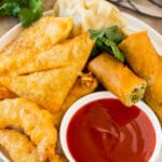 A bowl of sweet and sour sauce served with egg rolls, shrimp and wontons.