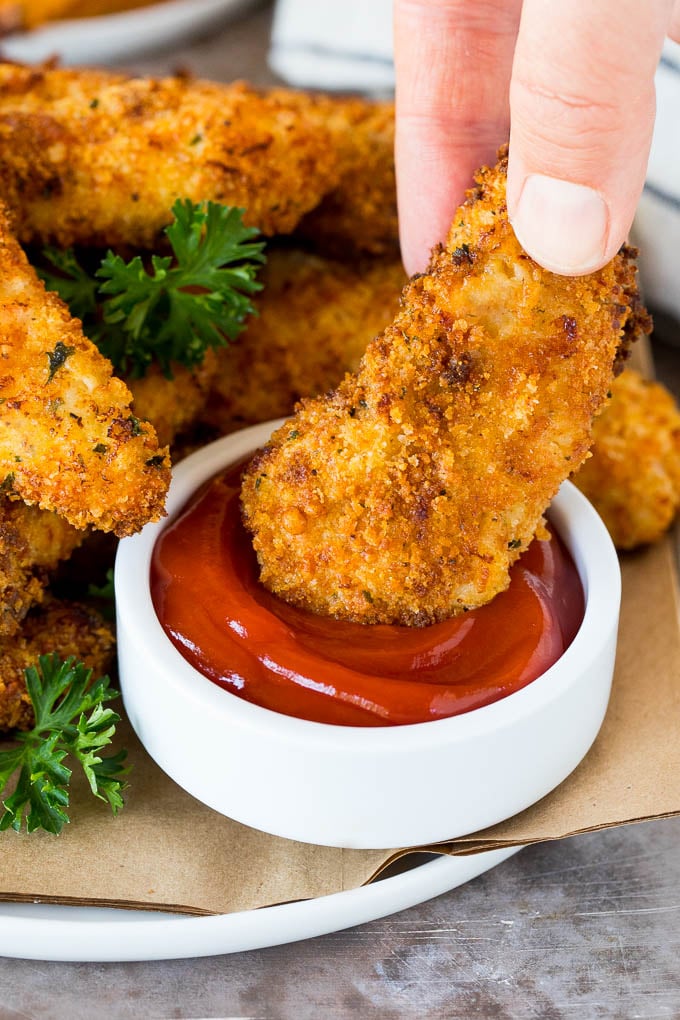 A hand dipping air fryer chicken tenders into a bowl of ketchup.