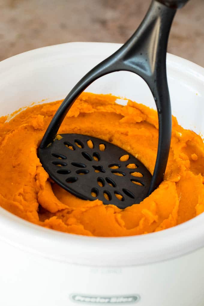 A potato masher that is mashing sweet potatoes in a crock pot.