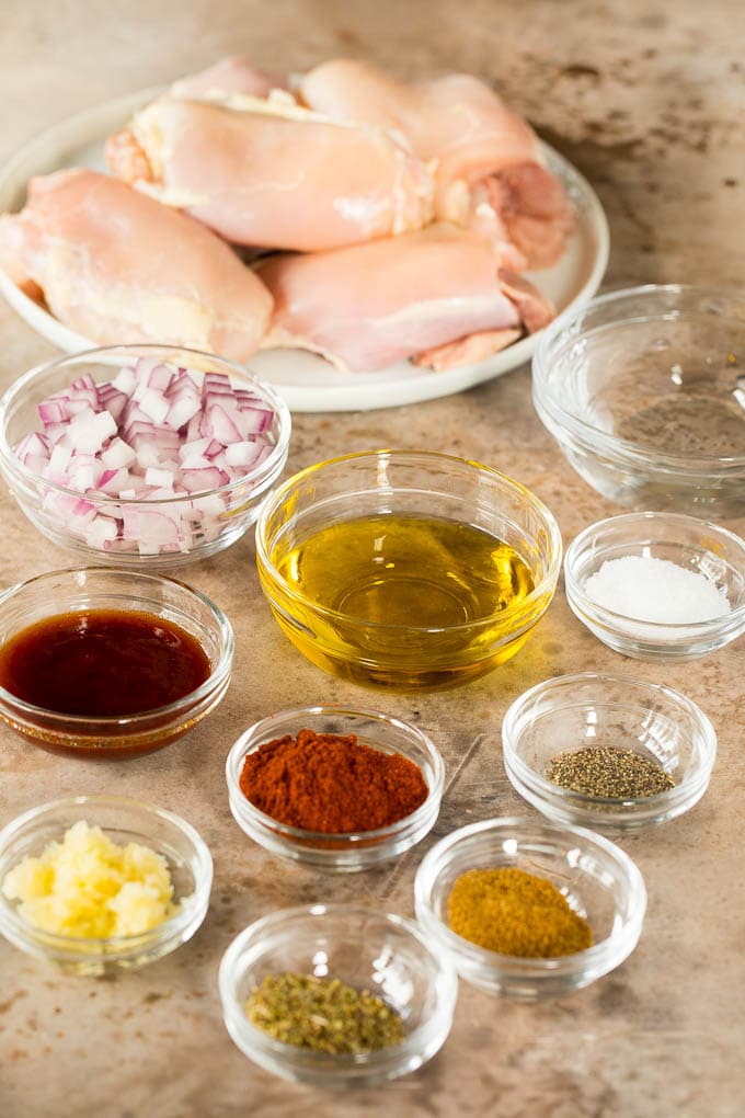 Chicken thighs with bowls of spices, olive oil, onion and garlic.