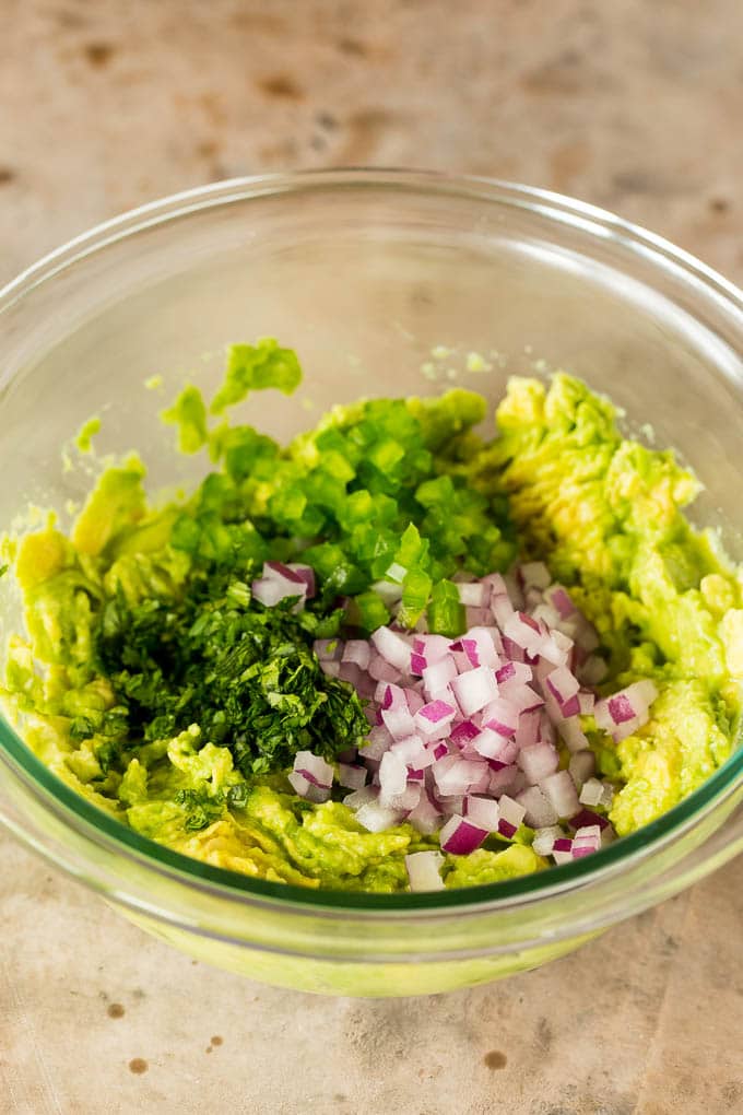 A bowl of mashed avocado with red onion, cilantro and jalapeno in it.