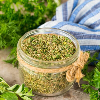 A jar of Greek seasoning surrounded by fresh herbs.