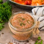 A jar of Creole seasoning surrounded by fresh herbs.