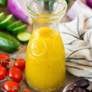 A bottle of Greek salad dressing surrounded by fresh vegetables.