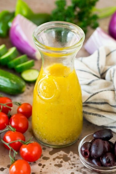 A bottle of Greek salad dressing surrounded by fresh vegetables.