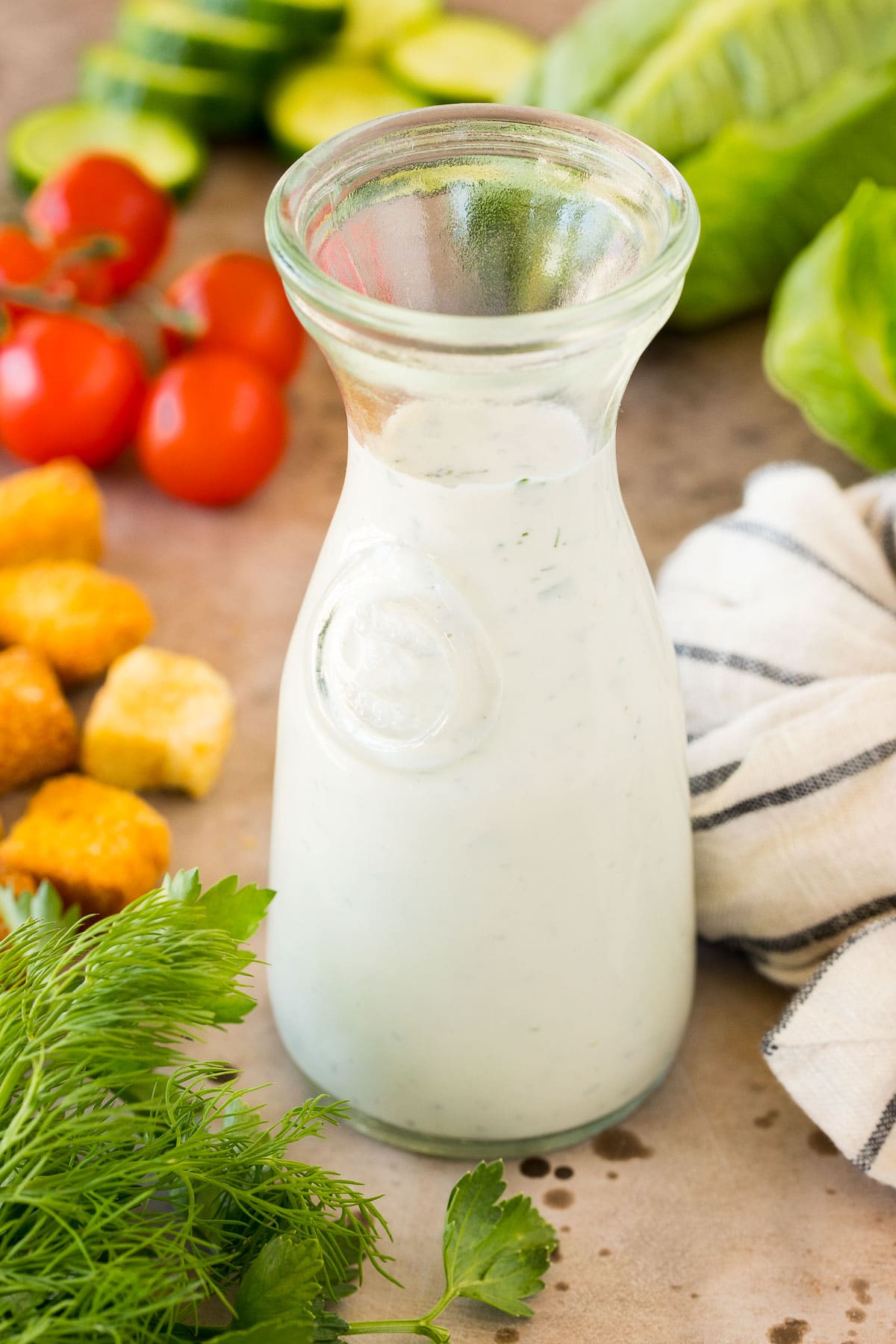 A glass container of homemade ranch dressing surrounded by salad ingredients.