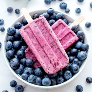 An image of two blueberry coconut popsicles in a bowl of blueberries.