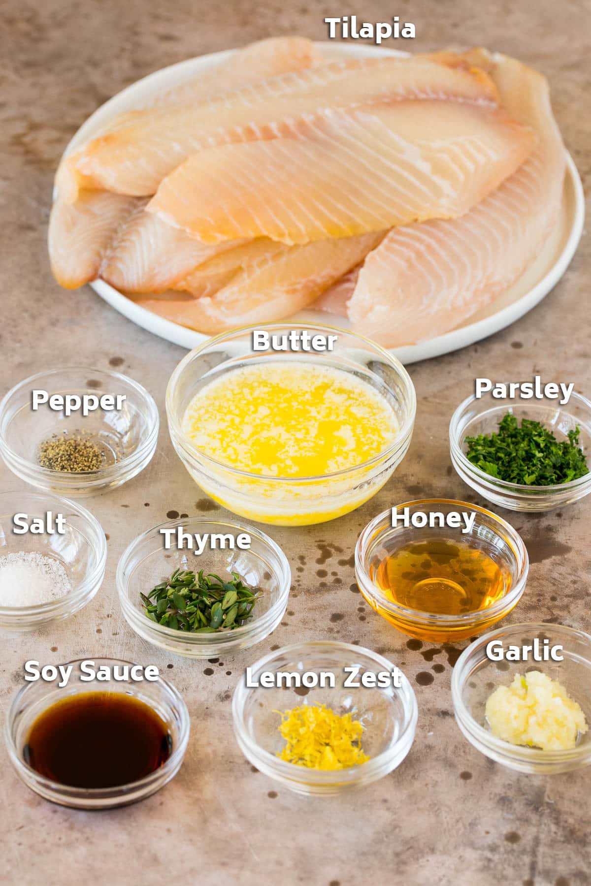 Fresh tilapia fillets with bowls of sauce ingredients including butter, lemon and herbs.