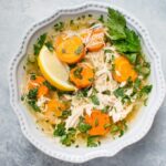 A bowl of lemon chicken orzo soup with carrots and a lemon wedge.