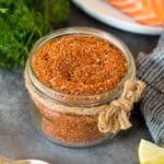 A jar of salmon seasoning with salmon, lemon wedges and parsley in the background.