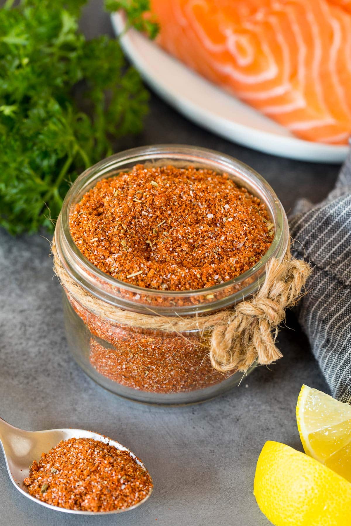 A jar of salmon seasoning with salmon, lemon wedges and parsley in the background.