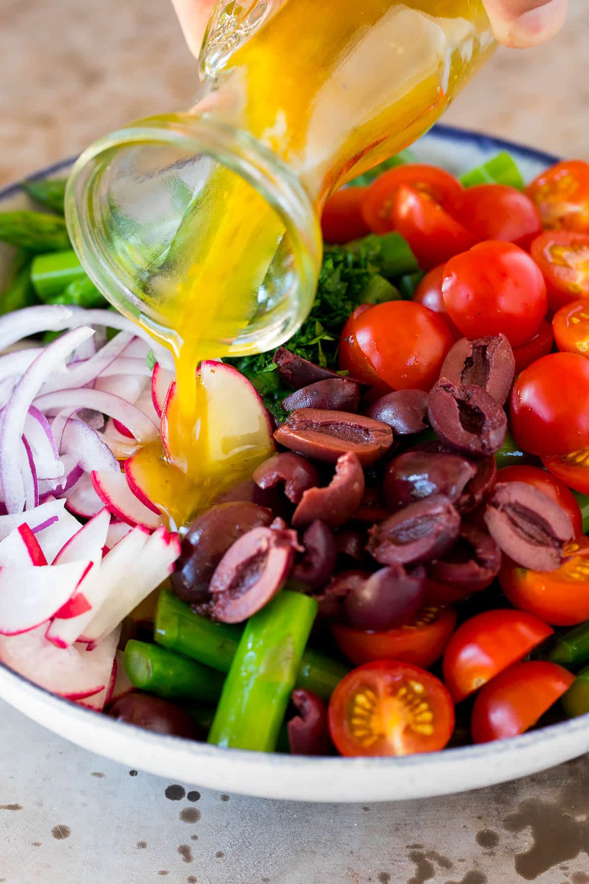 Dressing being poured over a bowl of vegetables and olives.