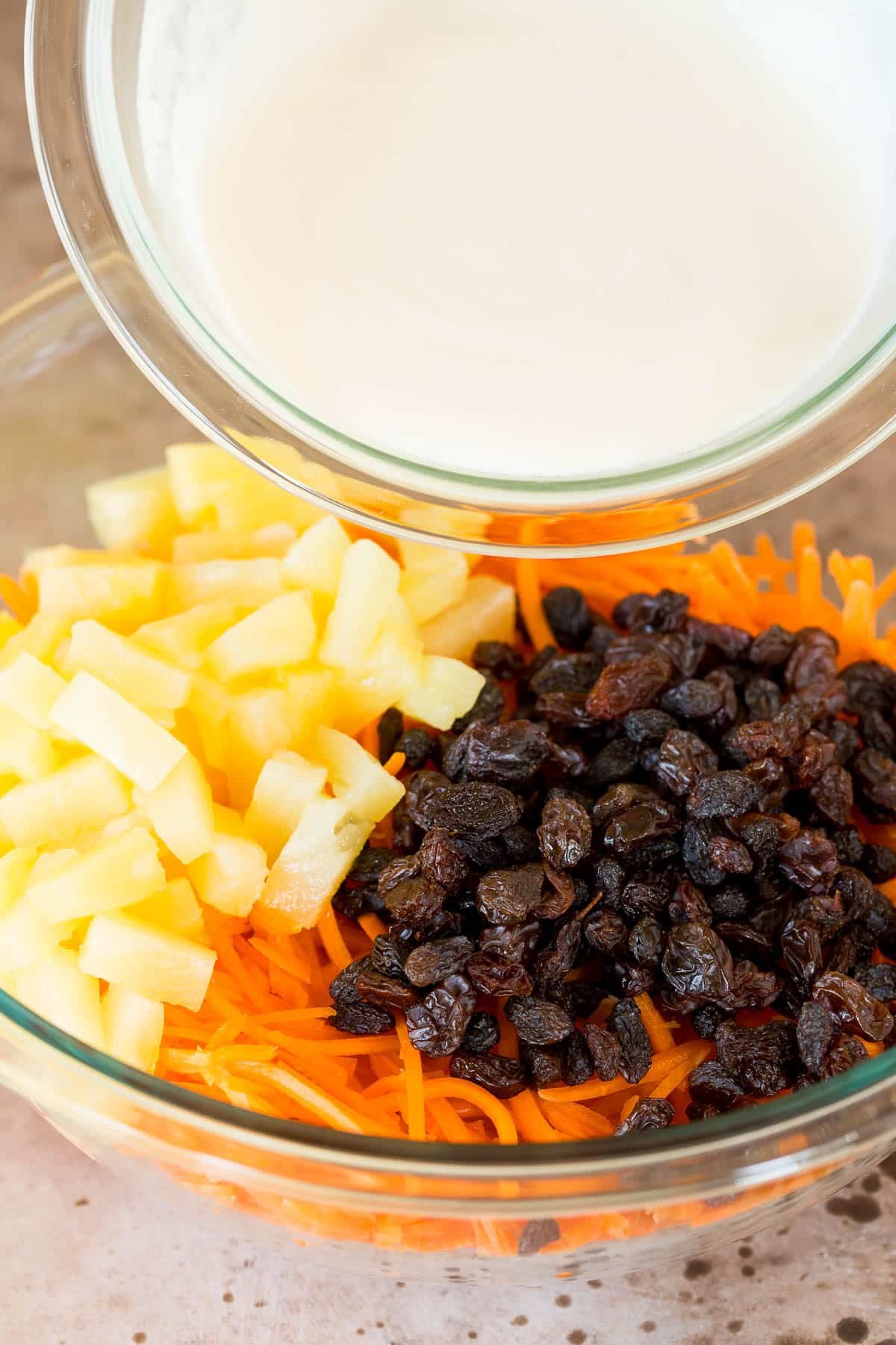 A bowl of dressing being poured over carrots, raisins and pineapple.