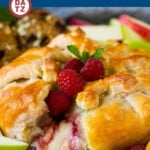 A baked brie on a plate which is brie topped with raspberry jam, fresh raspberries and candied pecans, all wrapped in puff pastry and baked.