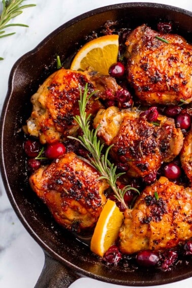 A skillet of cranberry chicken thighs with fresh cranberries, orange and rosemary.