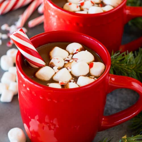 Mugs of peppermint hot chocolate topped with marshmallows and a peppermint stick.