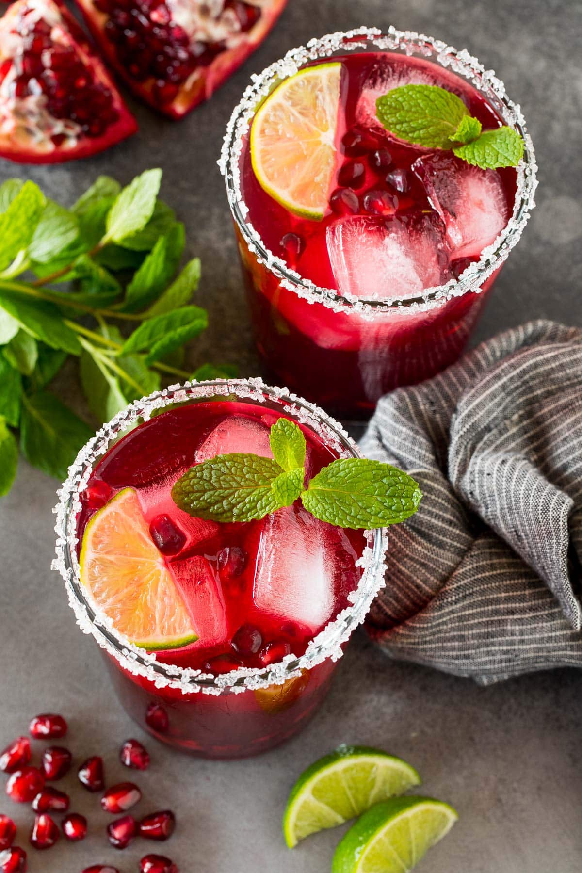 Two glasses of pomegranate margarita with lime slices, mint sprigs and fresh pomegranate seeds in them.