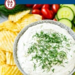 A bowl of dill dip surrounded by potato chips, carrots, cucumber slices and cherry tomatoes.