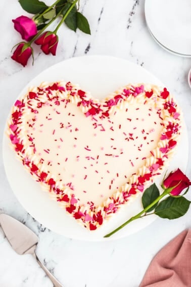 A heart cake on a cake stand topped with cream cheese frosting and red and pink sprinkles.