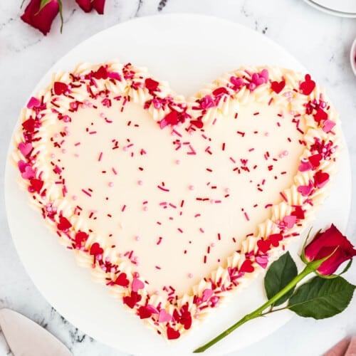 A heart cake on a cake stand topped with cream cheese frosting and red and pink sprinkles.